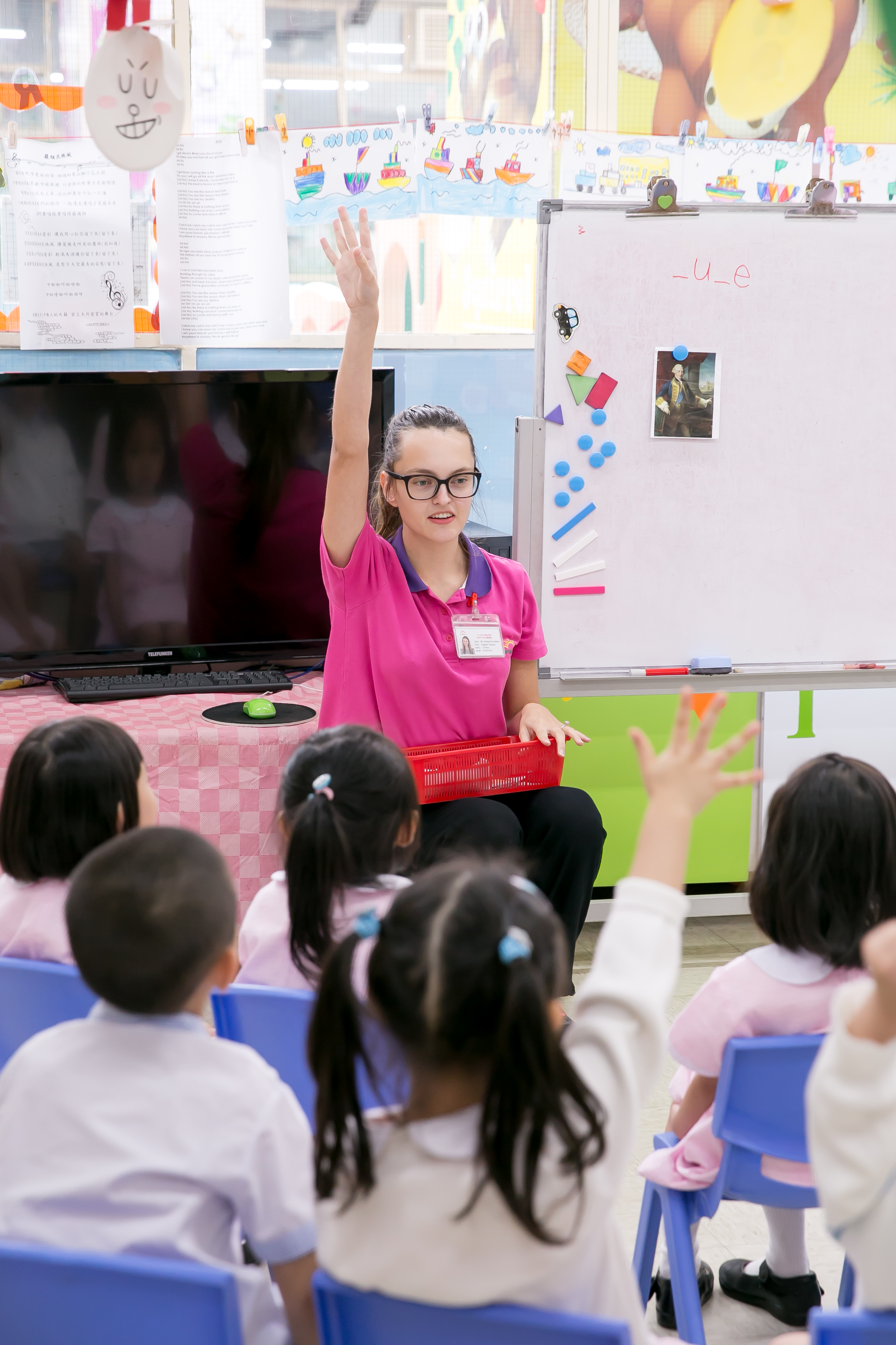 an early career TEFL teacher is sitting in front of a group of asian kindergarteners with her hand raised