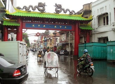 a chinese-style gate with grey dragons and people with wagons on the street of Surabaya, Indonesia after rain